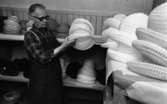 Hattfabrik läggs ned 1 mars 1966

En arbetare på en hattfabrik håller tre vita hattar i sina händer. Intill honom står hyllor fyllda med hattar.