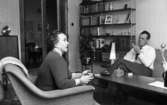 Brudpar 1946 (hemma hos), 9 mars 1966

En kvinna sitter i en fåtölj i ett vardagsrum. Hennes man sitter i en soffa mittemot. Mellan dem står ett bord med bl.a. ett askfat samt en glasskål med lock på. Båda personerna röker. I bakgrunden syns en bokhylla.