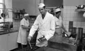 Fabriksmat, 9 mars 1966

En kock i vita arbetskläder lägger upp köttbullar i en förvaringslåda. I bakgrunden står två kvinnor och arbetar. Också de är klädda i vita arbetskläder.