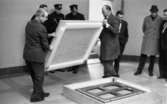 Nationalmuseum 1 31 maj 1966

Två män håller i ett stort trälock som de har lyft av en stor låda som ligger på golvet. En av männen bär en ljus arbetsrock. Två poliser står och tittar på i bakgrunden. Fyra andra herrar står också och betraktar verksamheten.