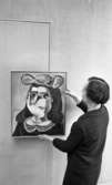 Nationalmuseum 1 31 maj 1966

En kvinna i mörk dräkt håller på att hänga upp en tavla på väggen. Den föreställer en kvinna.