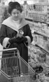 Burkmat 24 februari 1966

En kvinna klädd i mössa med tofs, kappa, tröja samt handskar betraktar en konservburk som hon håller i sina händer. Hon har en kundvagn med sig. Hennes handväska samt varor står i kundvagnen. Hon står intill hyllor fyllda med varor: paket samt konservburkar.