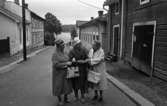 Noramarknad, Simmar i Gustavsvik 4 september 1965 

Tre äldre damer står på en gata i Nora. De är klädda i kappor, två bär hattar och alla bär handväskor i sina händer. Byggnader syns i bakgrunden.