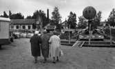 Noramarknad, Simmar i Gustavsvik 4 september 1965 

Tre äldre damer med ryggarna mot kameran går omkring på nöjesfältet på Noramarknaden. Två av dem är klädda i hatt och alla är klädda i kappor. En handväska syns på bilden. Flera åkattraktioner samt byggnader syns i bakgrunden.