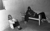 Oläslig rubrik 23 augusti 1965

En tonårsflicka sitter på en vit soffa och har en tonårspojkes huvud i sitt knä. En annan tonårsflicka sitter på golvet.