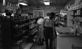 Persiko kvig, Laxar i Glanshammar 28 augusti 1965
 
En man står i förgrunden i en affär och betraktar en låda med persikor som ligger ovanpå en frysbox. I bakgrunden syns andra personer samt hyllor fyllda med olika varor.
