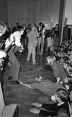 Popgala i Kumla, Farlig skolväg 24 augusti 1965

Ett rockband spelar på en scen i Kumla. Två av medlemmarna har gitarrer i händerna. En tredje har en tamburin i händerna. Sångaren till höger har en Sherlock Holmesinspirerad hatt på huvudet. Mikrofonstativ står på scenen. Ett piano syns i bakgrunden. Nedanför scenen står publiken. En tonårstjej ur publiken klättrar upp på scenen och tar tag i en av gitarristernas högra fot.