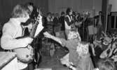 Popgala i Kumla,  24 augusti 1965

Ett rockband spelar på en scen i Kumla. Tre gitarrister syns på scenen varav en med en Sherlock Holmesinspirerad hatt på huvudet. En sångare står bredvid honom och sjunger i en mikrofon. Trummor samt ett piano syns i bakgrunden. Publiken står nedanför scenen. En tonårsflicka ur publiken sträcker ut sin vänstra hand mot en av gitarristerna.
