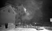 Branden under natten, 9 februari 1966

Ett hus brinner en vinternatt i februari. Rök väller ut ur en byggnad. Tre personer står på marken i förgrunden och betraktar scenariot. Det ligger fullt med snö på marken.