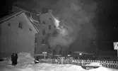 Branden under natten, 9 februari 1966

Ett hus brinner en vinternatt i februari. Rök väller ut ur en byggnad. Tre personer står på marken i förgrunden och betraktar scenariot. Det ligger fullt med snö på marken.