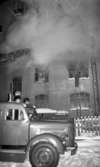 Branden under natten 9 februari 1966

I förgrunden syns en brandbil som står parkerad utanför en byggnad som brinner. Rök ses välla ut ur den och lutad mot husfasaden står en stege. På denna har en brandman klättrat upp. Han står strax utanför ett fönster på andra våningen.