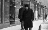 Brr va kallt, 4 februari 1966

En äldre herre klädd i hatt, rock, halsduk, byxor samt med handskar i vänsterhanden går gatan fram i centrala Örebro en vinterdag. I sin högra hand håller mannen en käpp. Snett bakom honom kommer en yngre man gående klädd i keps, hatt, rock, halsduk samt med galoscher på fötterna. Bakom herrarna syns skyltfönster med bl.a. damhattar i. Längre bort hänger en skylt vid sidan om en affär med texten 