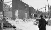 Byggspecial Oxhagen 11 februari 1966

En man står i överrock och hatt framför byggnader i området Oxhagen som håller på att byggas. Han har en portfölj under vänster arm. Två lyftkranar syns i bakgrunden på den snötäckta marken. Brädor ligger staplade under presenningar.