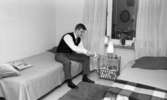 Byggspecial Oxhagen 11 februari 1966

En man sitter på en säng i Oxhagen och drar upp en väckarklocka. Han är klädd i vit skjorta, svart slips, grå byxor, svarta strumpor och svarta tofflor. I närheten av mannen finns en litet nattduksbord med en lampa och ytterligare en väckarklocka på. På sängarna ligger filtar och kuddar. I bakgrunden finns ett fönster med randiga gardiner.