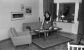 Byggspecial Oxhagen 11 februari 1966

I ett vardagsrum i en lägenhet i Oxhagen står en kvinna klädd i svart linne, grå kjol och sandaler och dukar ett bord. I rummet finns en soffa, en fåtölj och en matta under bordet.