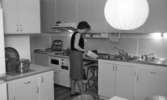 Byggspecial Oxhagen 11 februari 1966I ett kök i en lägenhet i Oxhagen står en kvinna och öppnar en påse som ligger på diskbänken. Hon är klädd i svart linne, grå kjol och sandaler.