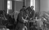 Rycker in i lumpen 8 februari 1966

Inne i ett stort rum håller fyra soldater på att klä på sig militäruniformer. Massor med kläder och utrustning ligger på bord och stolar som finns i lokalen.