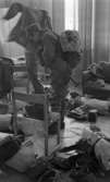 Rycket in i lumpen 8 februari 1966

Två soldater håller på att klä på sig militärkläder inne i en lokal. En soffa och en stolar finns också inne i lokalen. I bakgrunden syns ett stort fönster. Militär utrustning ligger runtomkring på en stol, på golvet och på soffan.