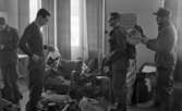 Rycket in i lumpen 8 februari 1966

Fyra soldater står inne i en stor sal och klär på sig militäruniform samt militärutrustning. Soldaten längst till höger håller på och hjälper sin kamrat till vänster med att knyta fast sovsäcken ovanpå ryggsäcken. Sovsäcken är ihoprullad.  Soldaten till vänster om dem håller i en påse med en sak i. På golvet står fullt med militär utrustning. Två fåtöljer, två stolar samt två fönster med gardiner kring syns i bakgrunden.