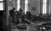 Rycket in i lumpen 8 februari 1966

Sex soldater står inne i en stor sal och klär på sig militäruniformer samt militärutrustning. I förgrunden ligger en stor låda som har packats ur. Militärutrustning ligger också på golvet samt på stolar runt omkring dem. Tre fönster med gardiner kring syns i bakgrunden.