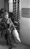 Rycket in i lumpen 8 februari 1966

En soldat är på väg ut genom en dörröppning samt ut ur en stor sal där han just har klätt om till militäruniform. Han bär en stor säck i sin vänstra hand. På dörren till höger står det 