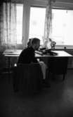 Student bostäder 1 februari 1966

En student i sin lägenhet sitter på en stol vid ett skrivbord. Bakom skrivbordet finns en glasdörr samt ett fönster. Ett antal pärmar ligger på bordet.
