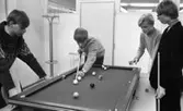 Biljard fritidssysselsättning 1 februari 1966

Fyra unga pojkar spelar biljard i ett rum. Pojken längst till vänster håller på att skjuta iväg en boll. De andra betraktar detta koncentrerat. Biljardbordet står ovanpå ett annat bord. Tre av pojkarna är klädda i tröjor och den fjärde bär kavaj. En klädhängare står i bakgrunden.