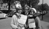 Skolavslutning 11 juni 1966

En tonårspojke står med cigarr i munnen och omfamnar två flickor i samma ålder under en skolavslutning. De håller gemensamt upp två betyg. Flickan till vänster har en hög hatt på huvudet och flickan till höger har en liten hatt på huvudet. Bilar står parkerade i bakgrunden.