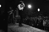 Popgala 6 juni 1966

En popgrupp uppträder på en scen i en stor hall. En sångare sjunger och en annan man spelar gitarr. Publik står framför scenen nere på golvet. Uppe på läktarna runtomkring sitter mer publik.
