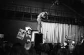 Popgala 6 juni 1966

En popgrupp uppträder på en scen i en stor hall. Sångaren står på en högtalare sjunger i en mikrofon. En musiker i bakgrunden spelar trummor. Publik står nere på golvet framför scenen.