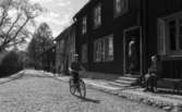 Wadköping, 28 maj 1966

En liten pojke kommer cyklande på en gata i Wadköping. Nära ett hus sitter en äldre dam klädd i klänning, kappa och hatt på en parkbänk. Fönstren på huset står vidöppna. En herre är på väg in i huset. ytterligare personer syns i bakgrunden.