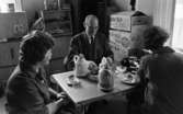 Lanthandel 9 juni 1966

En man och två kvinnor sitter vid ett fikabord under en kafferast i en lanthandel. De är klädda i arbetskläder. På bordet står två kaffekannor, en korg med fikabröd, tre kaffekoppar med fat och teskedar, en sockerskål samt en telefon. Bananlådekartonger står staplade i bakgrunden.