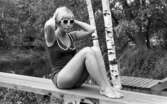 Baddräkter 30 juni 1966

En fotomodell klädd i baddräkt och solglasögon sitter på en träplanka. Hon är utomhus. I bakgrunden syns två björkar en brygga och ett vattendrag.
