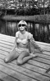 Baddräkter 30 juni 1966

En fotomodell klädd i bikini med volanger på och solglasögon sitter utomhus på en brygga. Ett vattendrag syns i bakgrunden.