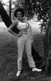 Baddräkter 30 juni 1966

En fotomodell klädd i vit top och vita byxor med svarta prickar på samt med vita stövlar på fötterna står och poserar  invid ett träd. Hon bär även solglasögon och har en kavaj i samma tyg som byxorna slängd över vänster axel.