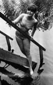 Baddräkter, Hus forts. 30 juni 1966

En fotomodell klädd i bikini med ett mönster med små stjärnor på står på en trappa som leder ner i ett vattendrag. I öronen har hon stora örhängen.