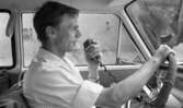 Beg. bilar, Ferieskola, Bodenkille slog rekord 14 juli 1966

En ung man sitter i framsätet i en vit bil och håller i ratten samt i en högtalarmikrofon. Han är klädd i vit skjorta.