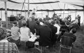Helgelseförbundet, Japan på Svampen 24 juni 1966

En predikant står inne i ett stort vitt tält och predikar. Han pekar med vänster hand upp mot himlen. Ett antal personer sitter på träbänkar och lyssnar på honom. En herre sitter på en fällstol.