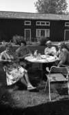 Lekhyttan 21 juni 1966

Två äldre damer och två äldre herrar sitter vid ett dukar runt litet bord utomhus och dricker kaffe i Lekeberg. Damerna är klädda i korta klänningar och en av dem har även ett förkläde på sig och herrarna är klädda i skjortor och byxor. Bordet är dukat med kaffekanna, kaffekoppar, bullfat etc. Ett stort hus syns i bakgrunden.