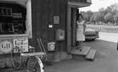 Lekhyttan 21 juni 1966

Närbild på en kvinna i kort, ärmlös sommarklänning som står på entrétrappan till Lekhyttans handel. En cykel står parkerad i cykelstället invid huset och en bil syns till höger i bakgrunden.