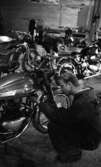 Motorcyklar, långtradare i diket 30 juni 1966En ung man i arbetskläder sitter på huk och servar en motorcykel på en verkstad. I bakgrunden syns flera motorcyklar.