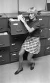 Kort- kort på jobb 2 augusti 1966

En ung kvinna i kortklippt ljust hår, kortärmad tröja, skotskrutig kjol och sommarskor står invid ett arkiv med en massa lådor. Hon bläddrar bland mapparna i en av lådorna.