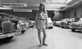 Kort- kort på jobb 2 augusti 1966

En ung kvinna med långt hår, långärmad tröja, kort kjol, halsband runt halsen och med sandaler på fötterna står inne i en lokal fylld med bilar. Två skyltar hänger i taket. På den ena står det: 