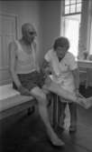 Loka brunn 22 juli 1966

En äldre herre klädd i vit nätundertröja samt mörka shorts sitter på en brits och sträcker ut vänstra benet. En kvinna klädd i vita arbetsklädsel håller i hans ben. Ett vitt bord syns i bakgrunden.