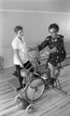 Loka brunn 22 juli 1966

En äldre dam i knäkort klänning cyklar på en motionscykel under överinseende av en arbetsklädd tränare på kurorten Loka brunn.