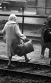 På stationen 1 16 augusti 1966

En äldre dam går över järnvägsspåren på Örebro station. Hon är klädd i ljus kappa, ljus hatt med genomskinlig sjalett över och ljusa tofflor på fötterna. Hon har två väskor. Hon bär en i den ena handen och en i den andra.