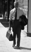 Strosor rep 2 25 juli 1966

En herre promenerar på trottoaren i centrala Örebro klädd i vit skjorta, svart slips, svarta byxor och svarta skor. Han håller en svart portfölj i sin högra hand. Slängd över hans vänstra arm är hans svarta kavaj.