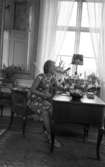 Svartå Herrgård 28 juli 1966

En kvinna i kort, ärmlös sommarklänning och sandaletter på fötterna sitter vid ett skrivbord i en salong på Svartå Herrgård. En vas med en blombukett i står på bordet. Ett fönster syns i bakgrunden.