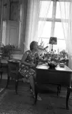 Svartå Herrgård 28 juli 1966

En kvinna i kort, ärmlös sommarklänning och sandaletter på fötterna sitter vid ett skrivbord i en salong på Svartå Herrgård. En vas med en blombukett i står på bordet. Ett fönster syns i bakgrunden.