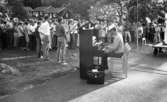 Missionsläger 10 augusti 1967

På ett missionsläger sitter en man och spelar piano utomhus. Han är klädd i vit hatt, mörka solglasögon, randig tröja, och mörka byxor. En stor, svart portfölj står på marken bredvid honom. Flera musiker med tromboner i sina händer står i närheten av honom samt flera sångare skymtar längre bort i publiken. Till höger syns en dirigent som står på ett podium med hjul.